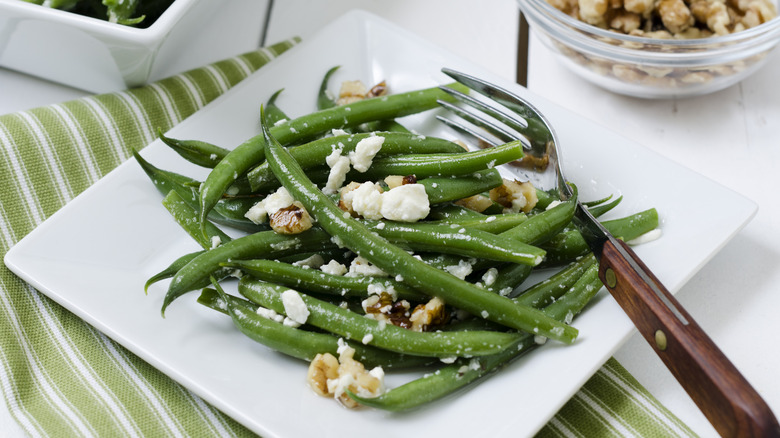 Green beans on a white plate with goat cheese crumbles and chopped walnuts.