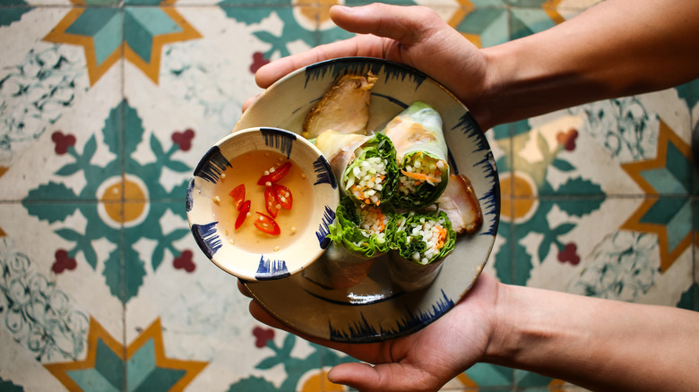 Two hands holding a bowl of summer rolls with nuoc cham