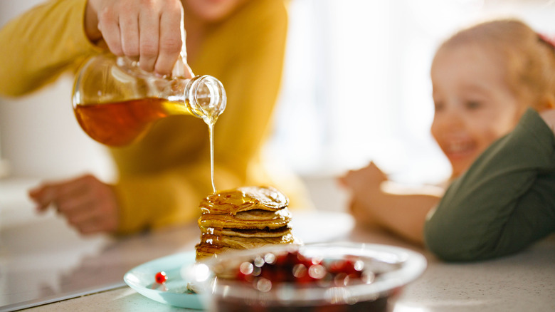 Mom drizzling pancakes over kids' stack of pancakes on plate