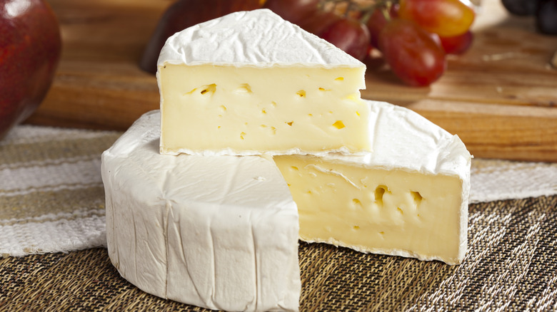 Wheel of Brie with a slice cut out and placed on top on a table with a cloth woven napkin and grapes in the background on a wooden board.