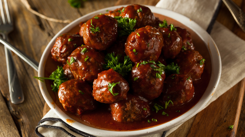 A bowl of meatballs sitting in a brownish-red sauce in a white bowl with bits of fresh-chopped parsley