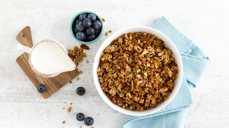 A bowl of granola with nuts next to a small pitcher of milk and a tiny bowl of blueberries