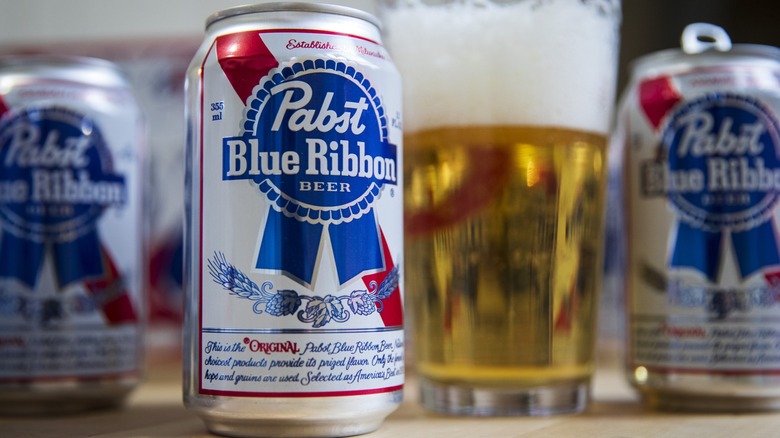 Cans of Pabst Blue Ribbon beer next to a glass of beer