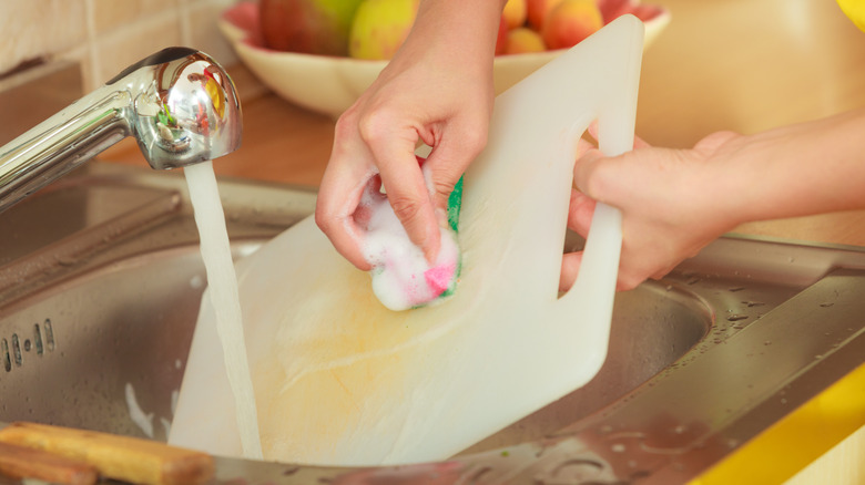 person scrubbing a plastic cutting board in a sink under a running faucet