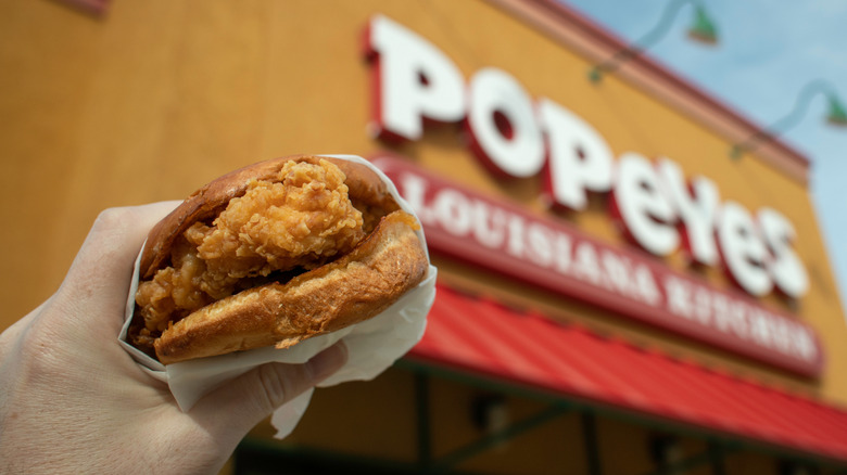 person holding up a Popeyes chicken sandwich with the exterior of the fast food restaurant in the background