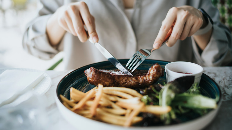 Diner using knife and fork to cut steak on a plate with fries and a salad