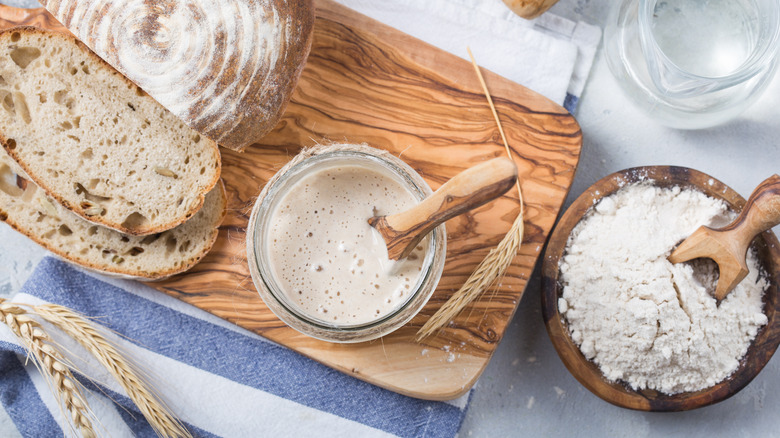 Overhead of sourdough starter in an open jar on a board with sliced sourdough and bowl of flour