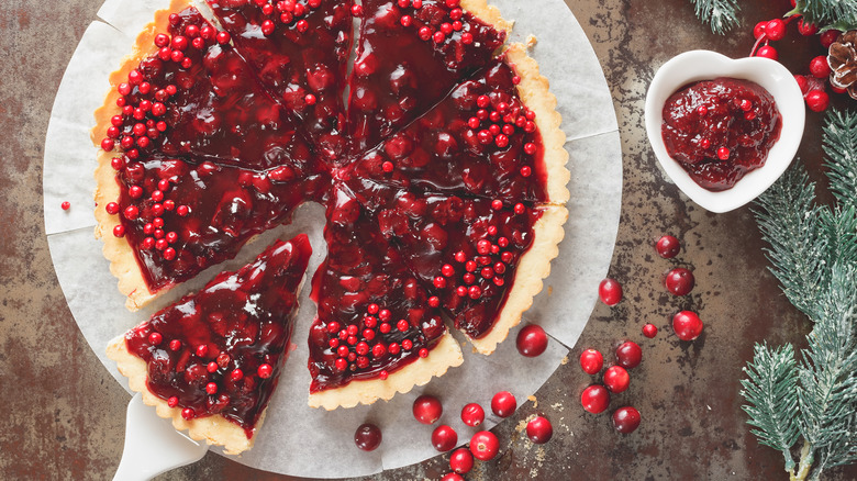 A large cranberry tart, neatly cut into eight slices, with a side of cranberry sauce and pine tree needles scattered to the right.