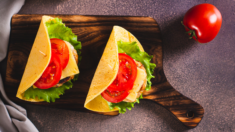 Two tacos on a wooden board filled with lettuce, meat, tomato, and cheese