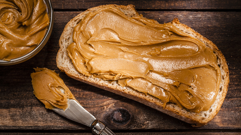 peanut butter spread on wheat bread with a bowl of peanut butter on a dark background