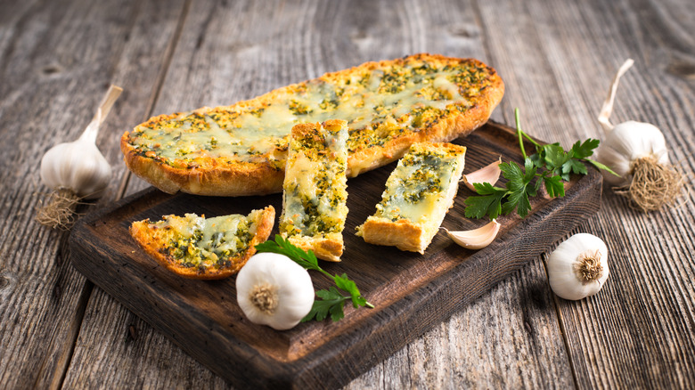 Garlic bread on board surrounded by cloves of garlic