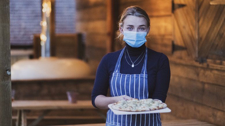 Waitress wearing a surgical mask looking at the camera and holding a pizza