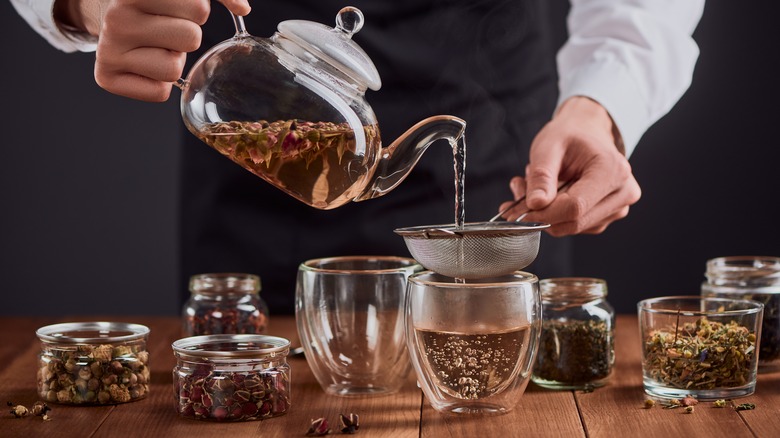 Person pouring loose leaf green tea from a glass teapot through a strainer into a glass tumbler