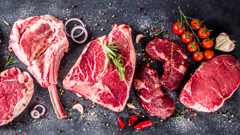 Various cuts of beef steaks laid out next to each other, adorned with vegetables, herbs, and spices