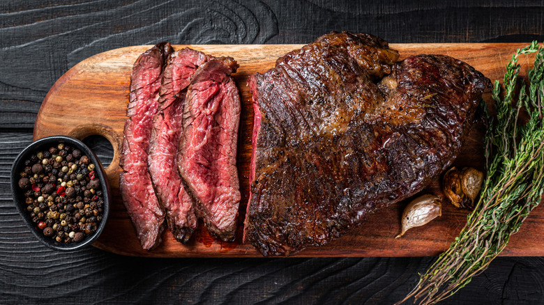 Grilled hanger steak on wood board with rosemary bunch and bowl of peppercorns