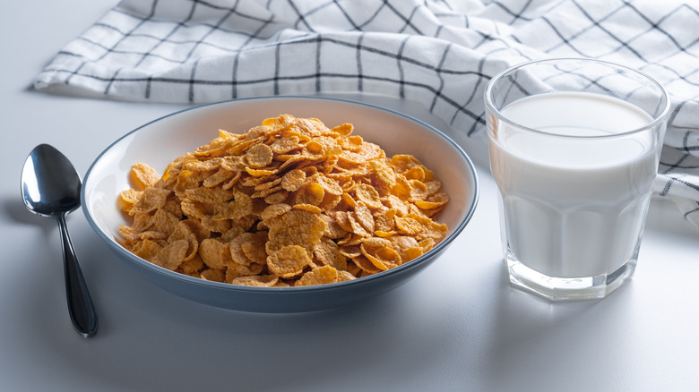 A dry bowl of corn flakes with a glass of milk to the side