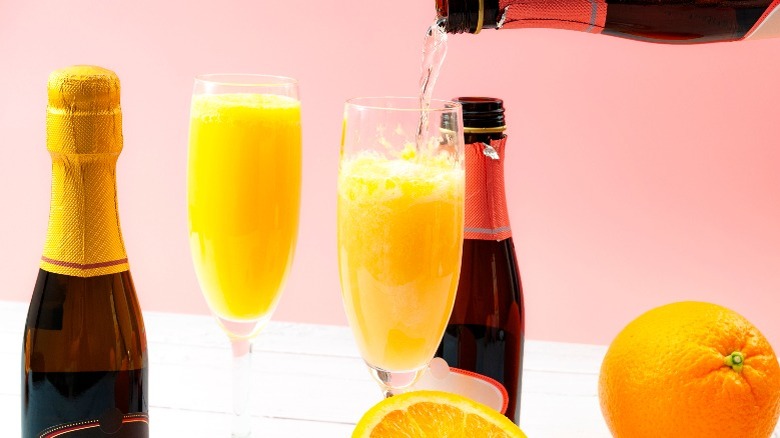 Mimosa pouring from a bottle into a flute next to Champagne bottles, a Mimosa, and oranges