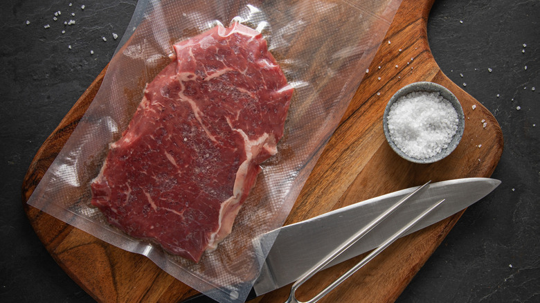 A large steak inside a vacuum-sealed bag for sous vide cooking, with a knife and sea salt