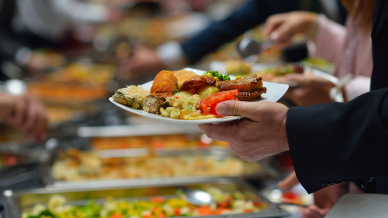 Hands holding a full plate of food in front of a busy buffet