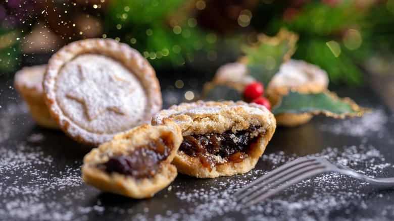 A small mincemeat pie, split open to show the gooey brown filling, with other pies in the background, dusted in powdered sugar