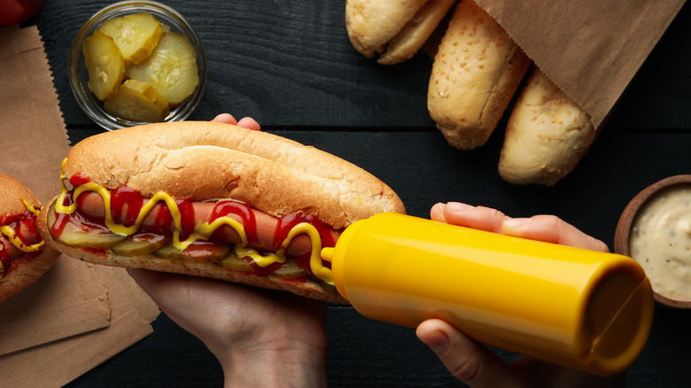 Person squeezing mustard on hot dog surrounded by ingredients