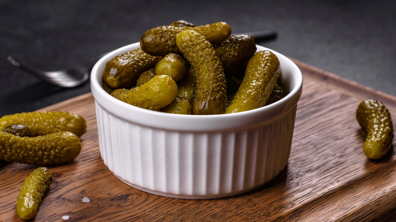 Ramekin full of small pickles, with pickles to either side on wooden board