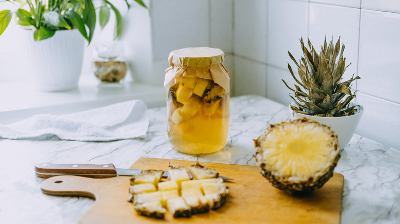 Covered jar of tepache with cutting board of sliced pineapple