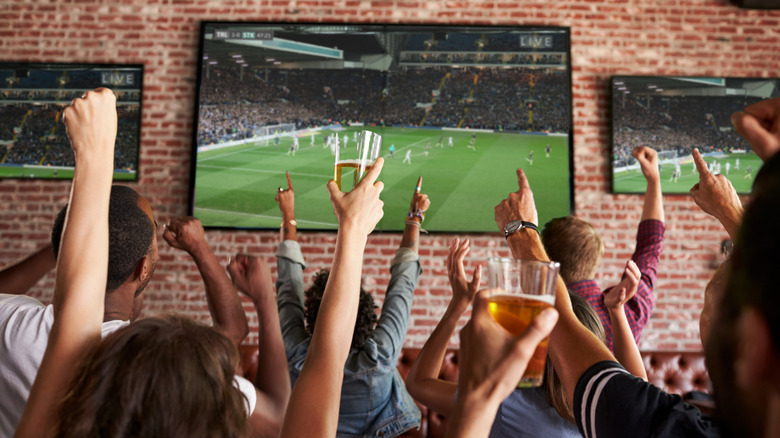 big screen TVs on the brick interior wall of a sports bar, with patrons cheering and raising their hands in the foreground