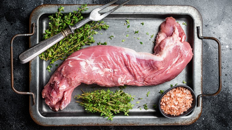 Raw pork tenderloin on baking sheet with fresh herbs and coarse sea salt