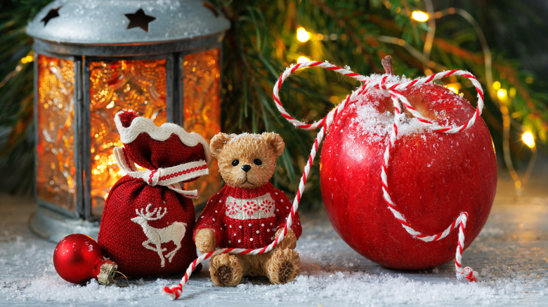 Christmas card with a red apple, teddy bear, gift bag and a lantern on a table.