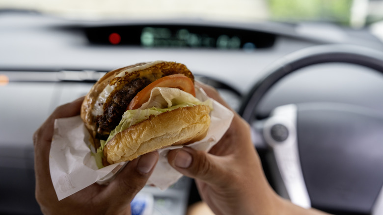 A close-up of an all-dressed, juicy burger in the hands of someone sitting in their car.