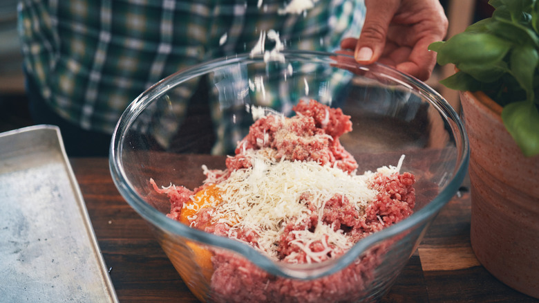 Chef adding grated cheese to meatball mixture in glass bowl