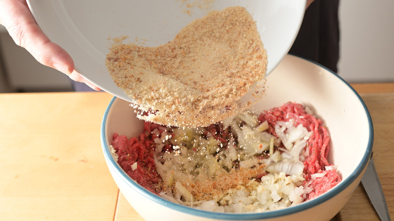 Breadcrumbs being poured into white bowl of ground meat