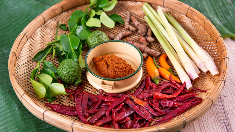 Ramekin of Thai red curry paste, surrounded with the ingredients used to make it: Limes and lime leaves, lemongrass, dried and fresh chilies, and galangal