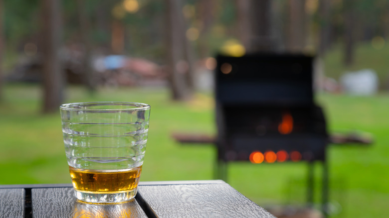 Glass of whiskey on picnic table edge with grill in background