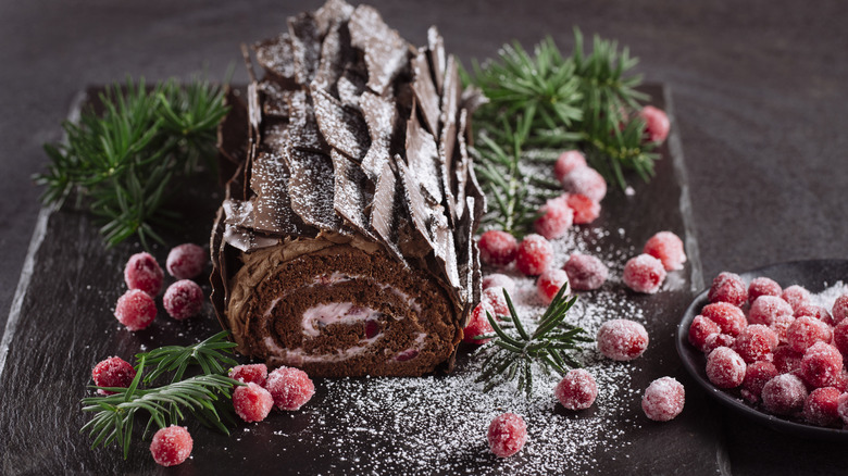 A creamy Yule log topped with sugared cranberries sits on a wooden table.