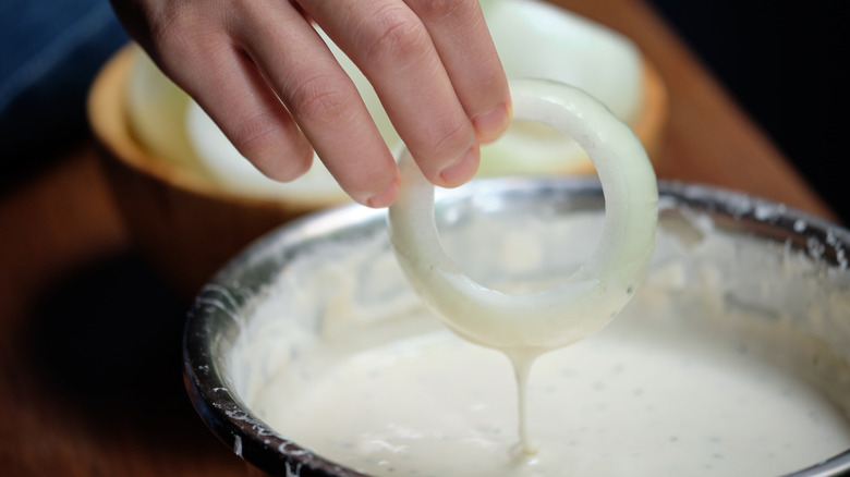 Hand dipping onion ring in bowl of batter.